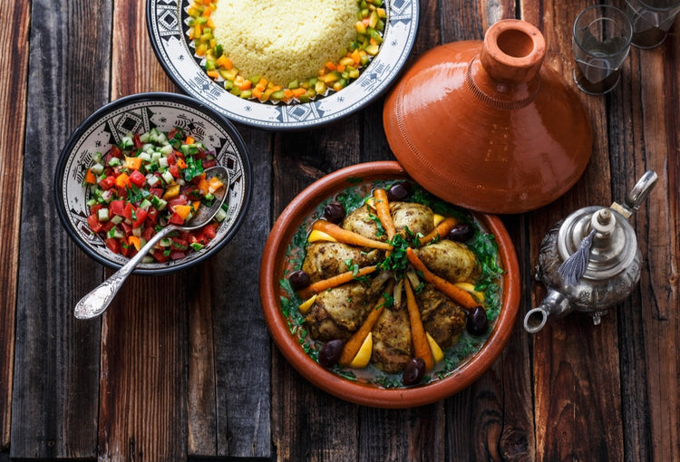 Vue aérienne d’un repas marocain traditionnel avec un tajine de poulet, un plat de couscous garni de légumes, une salade fraîche aux tomates, concombres et oignons, le tout disposé sur une table en bois avec une théière orientale.